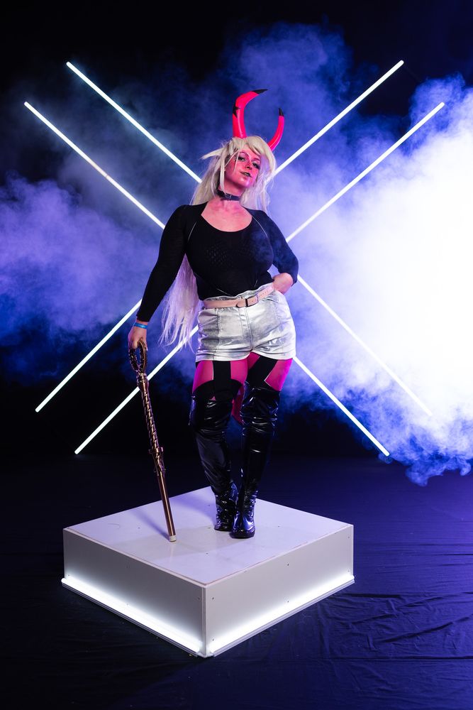 Professionnal cosplay picture of Bilie as Verosika from Helluva Boss. She is posing with a fancy backdrop with a proud and seductive pose
