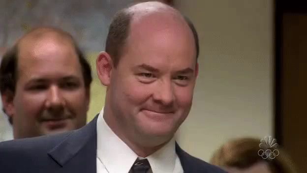 a bald man in a suit and tie is smiling while standing in front of a group of bald men .