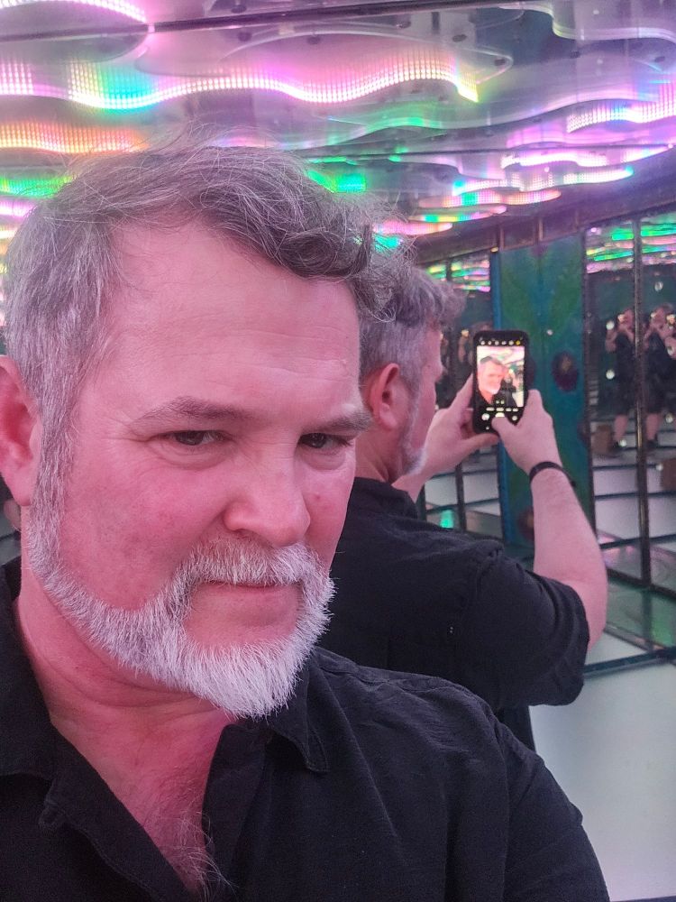 Overly Florida fifty-something guy with gray beard and black shirt in mirrored room so the selfie-camera is visible behind him.