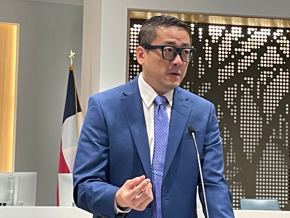 Photo of a man in a blue suit and tie and fantastic glasses speaking at a podium in the Commissioners’ courtroom. 
