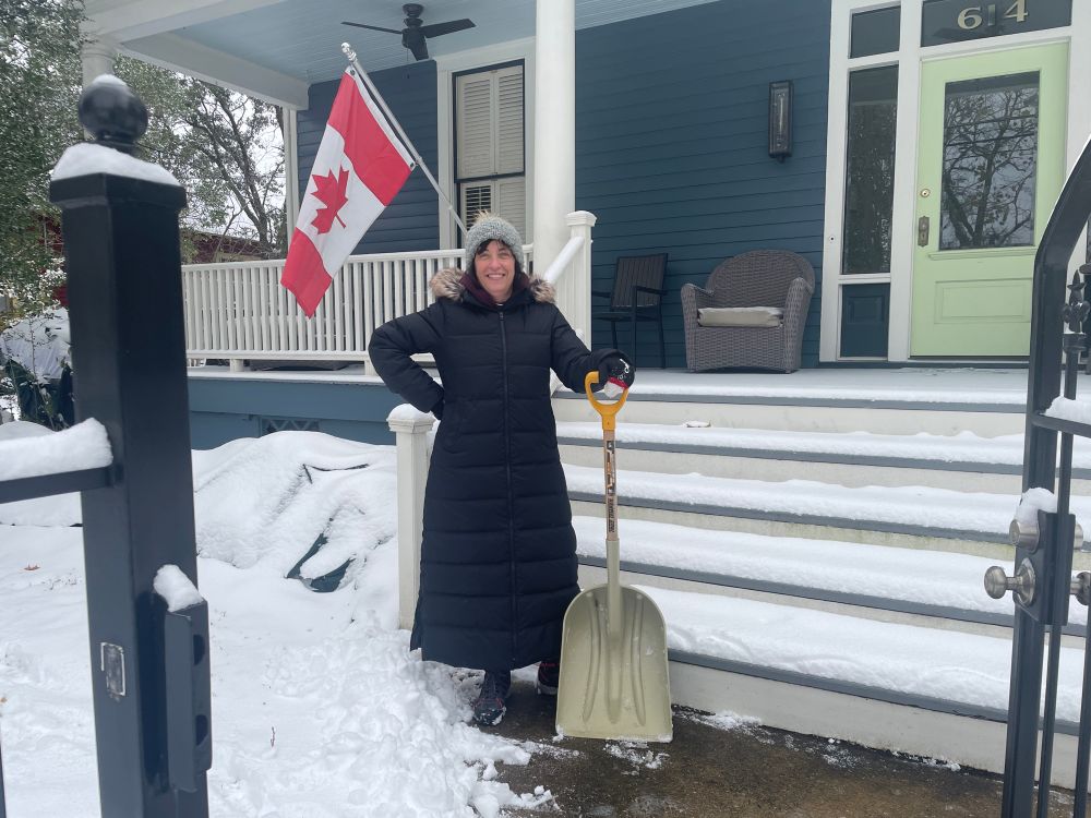 Photo of a woman with a shovel and cleared front walk in front of her house. She holds a snow shovel and a Canadian flag flies over her shoulder, so you know she knows snow. She was about to shovel the front stairs. 