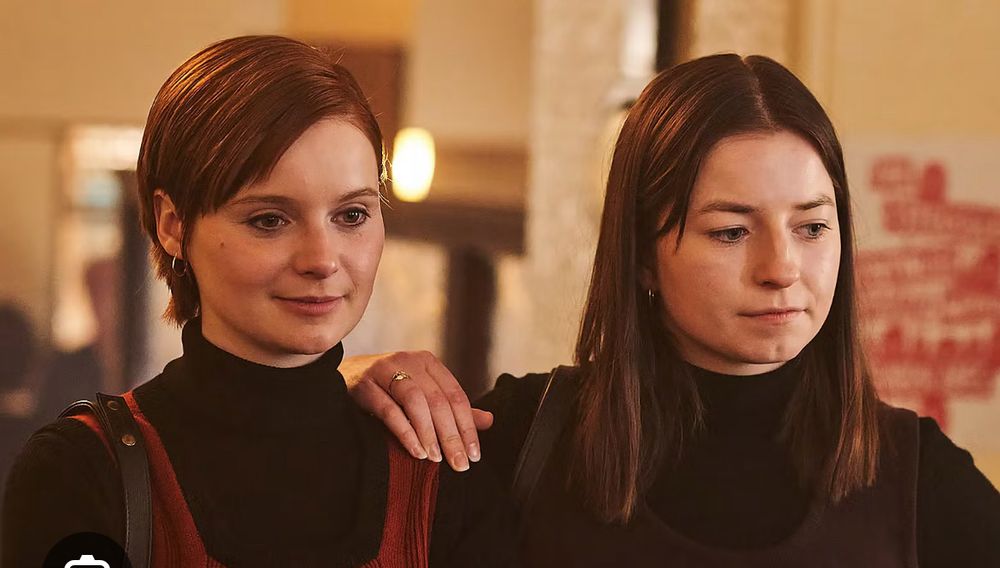 Two women standing together looking at something in front of them. The one on the left has pale white skin, slay check, and straight red hair in a longer pixie cut. She wears a black turtleneck and deep red pinafore over it. On the right the woman, also with pale white skin, has long straight warm brown hair.  She has on a black turtleneck as well. They are the actresses playing sisters Delours and Marian Price in Say Nothing, a docu-drama about members of the IRA. 