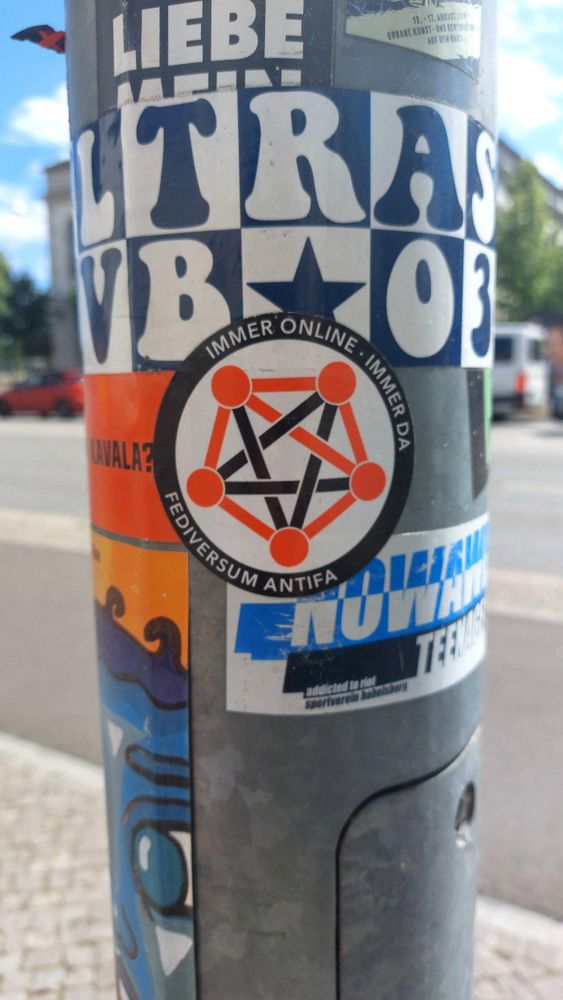 Foto eine Laternenmastes mit vielen Aufklebern, zentral ein runder Aufkleber mit fünf roten, im Kreis angeordneten Punkten, die alle paarweise mi dicken roten und schwarzen Linien verbunden sind. Darum herum ein schwarzer Rand mit weißem Text: "Immer online - immer da / Fediversum Antifa"