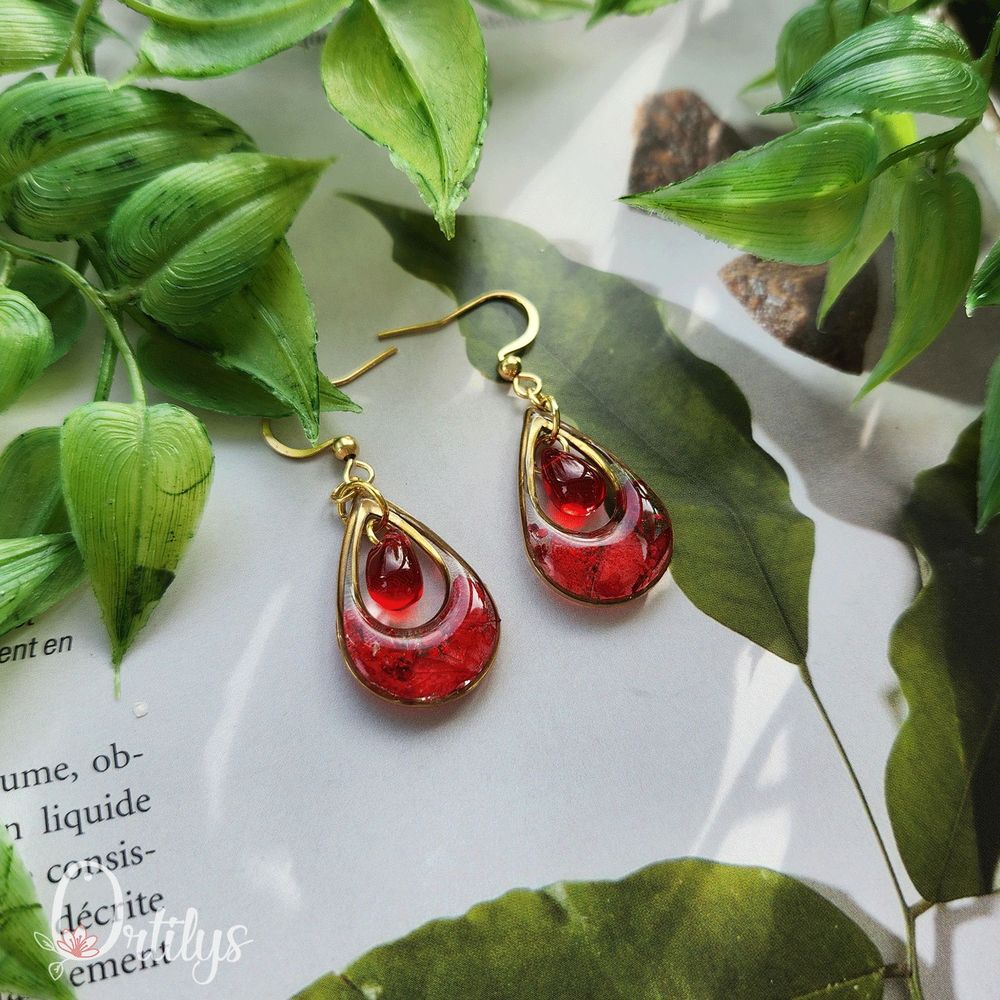 Des boucles d'oreilles en forme de goutte, dont le centre est creux pour y accueillir une perle en verre. Ici, les boucles sont dorées, les fleurs du contour sont rouges et la perle est rouge transparente