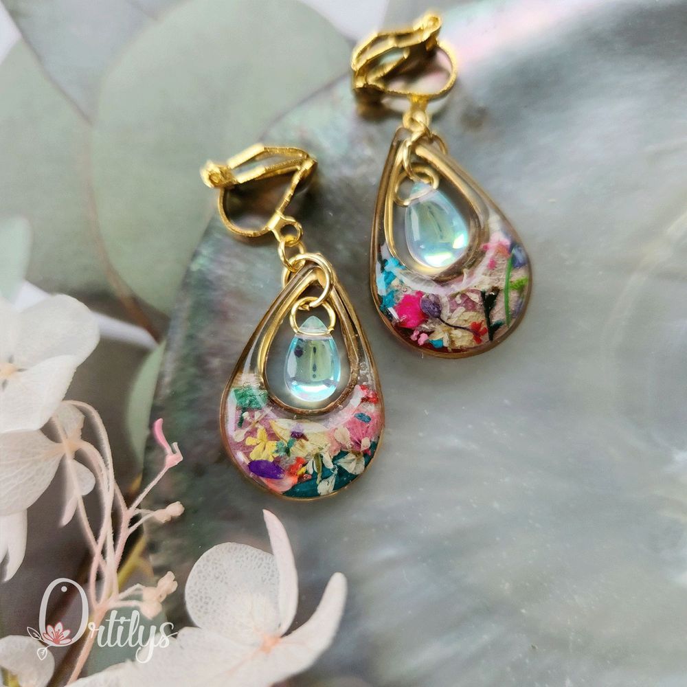 Des boucles d'oreilles en forme de goutte, dont le centre est creux pour y accueillir une perle en verre. Ici, les boucles sont dorées, les fleurs du contour sont multicolores et la perle est couleur diamant