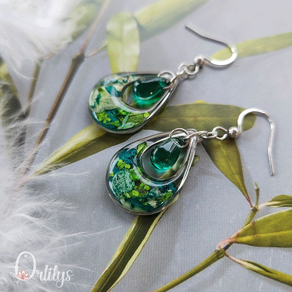 Des boucles d'oreilles en forme de goutte, dont le centre est creux pour y accueillir une perle en verre. Ici, les boucles sont argentées, les fleurs du contour sont de différents verts et la perle est vert émeraude