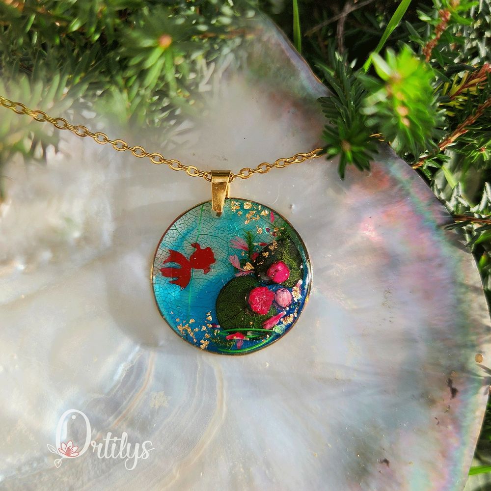 Pendentif doré  rond en résine avec fleurs séchées roses et feuillages verts, ainsi qu'une petite carpe rouge en résine ondulant à l'intérieur, inspiré d’un bassin de nymphéas