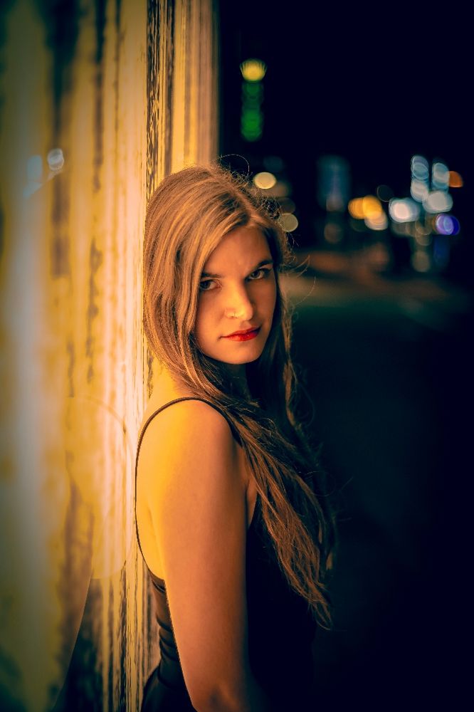 Das Bild zeigt eine junge Frau bei Nacht. Sie hat langes, gewelltes, blondes bis braunes Haar, das über ihre linke Schulter fällt. Sie schaut direkt in die Kamera.  Ihre Lippen sind mit rotem Lippenstift geschminkt. Der Hintergrund ist unscharf und zeigt eine Straße mit verschwommenen Lichtern, die eine nächtliche Atmosphäre andeuten. Die Frau lehnt an einer strukturierten, hellgelben oder goldenen Wand. Das Foto ist von der Seite aufgenommen und die Beleuchtung kommt von vorne und von der Seite, was einen schönen Kontrast zwischen der Frau und dem Hintergrund schafft.
