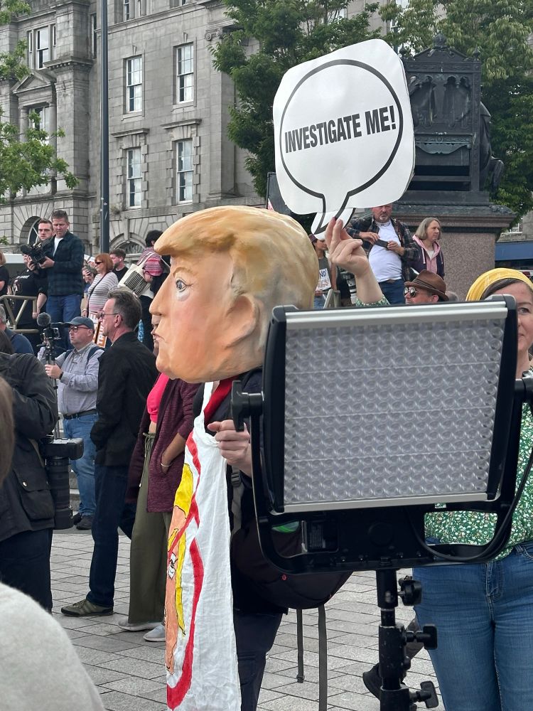 Protesters at anti-Trump rally. One person is wearing an oversized model of Trump’s head with a sign saying ‘investigate me!’