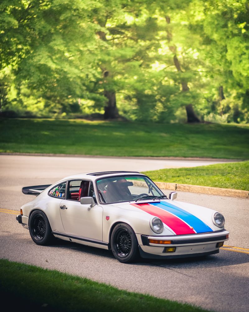 a white Porsche 930 with red and blue racing stripes on the hood and a collection of stickers on the rear side window, in the sun, driving along a road next to a forest