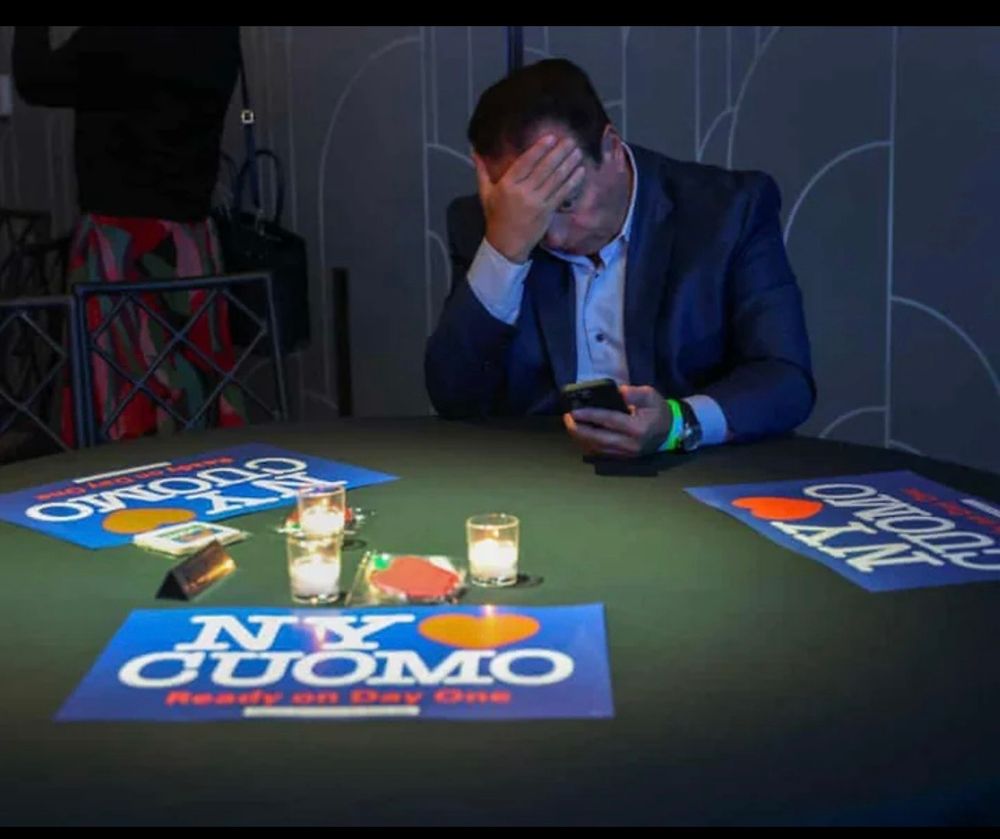 cuomo in his feelings after losing the election on November 4. A table with New York loves Cuomo signs a few drinks and Cuomo with his hand on his forehead, looking down to his phone, sad and alone. 
