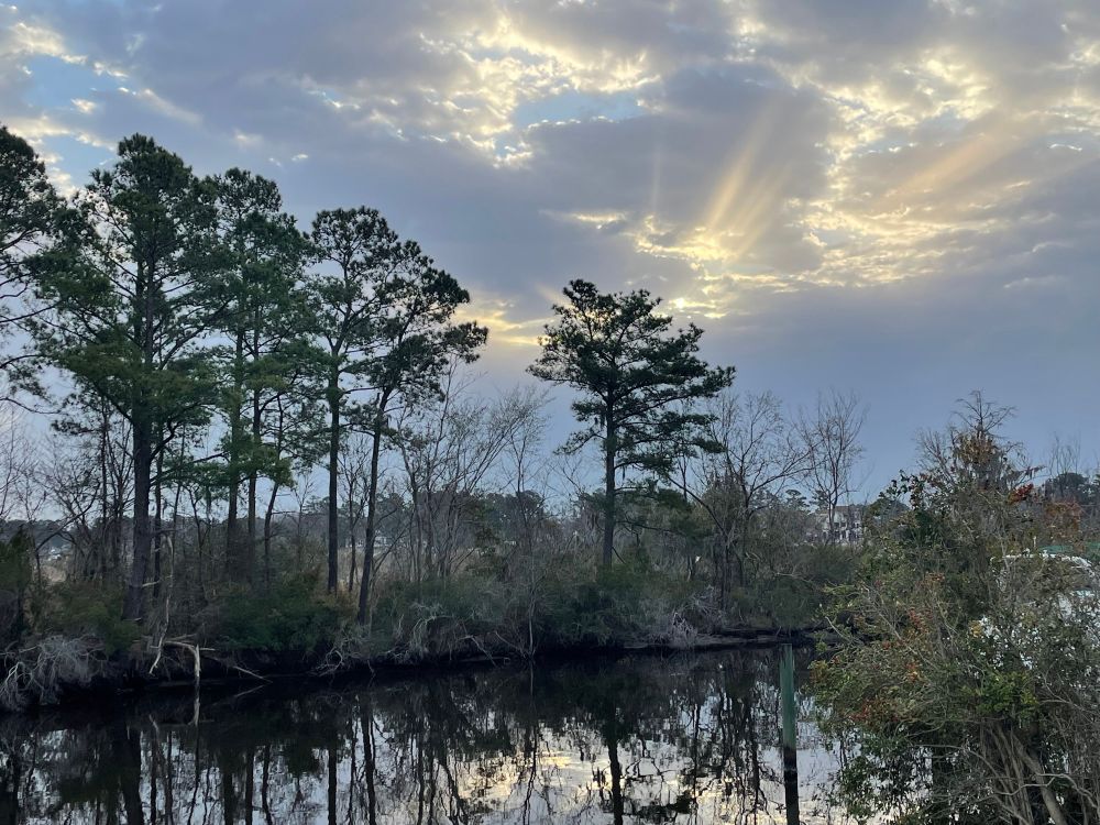A river, smooth, with the sky reflecting off it. There is cloud cover with some sun rays poking through. It’s peaceful.
