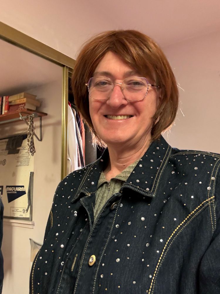 Woman with reddish brown hair, gold framed glasses with pink highlights, pink star-shaped earrings, and wearing a dark blue denim jacket with rhinestones over a gray ribbed sweater. She is smiling broadly because she is finally able to live as her true self after sixty years.