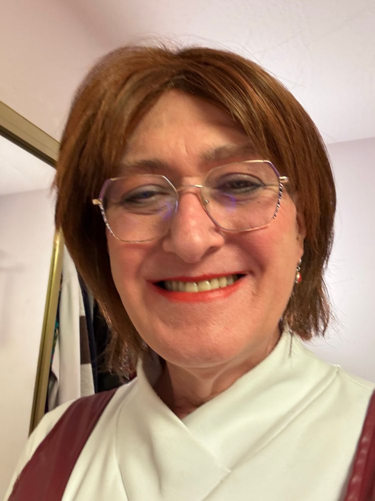 Smiling woman with reddish brown hair, hazel eyes, gold frame glasses, red lipstick, and dangly red earrings.