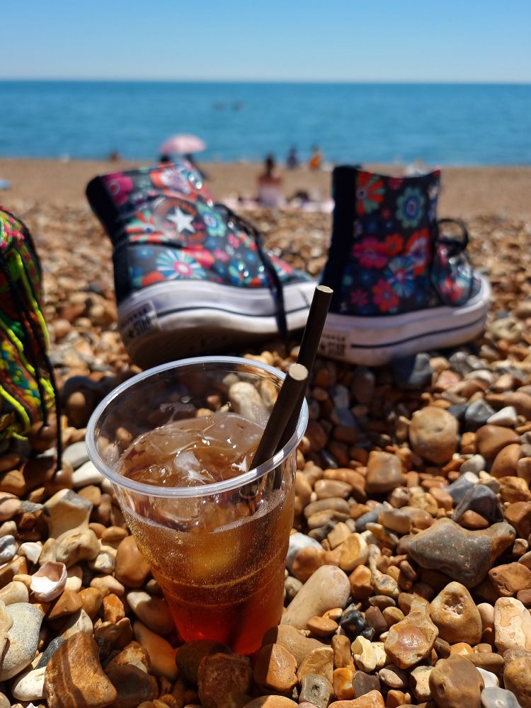 A pair of floral converse shoes and a cup of iced tea on the pebbled Brighton beach, with the sea in the background.