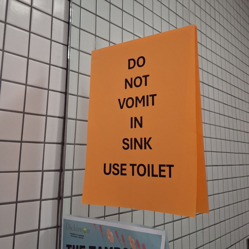 Orange sign on mirror that says, "Do not vomit in sink. Use toilet."