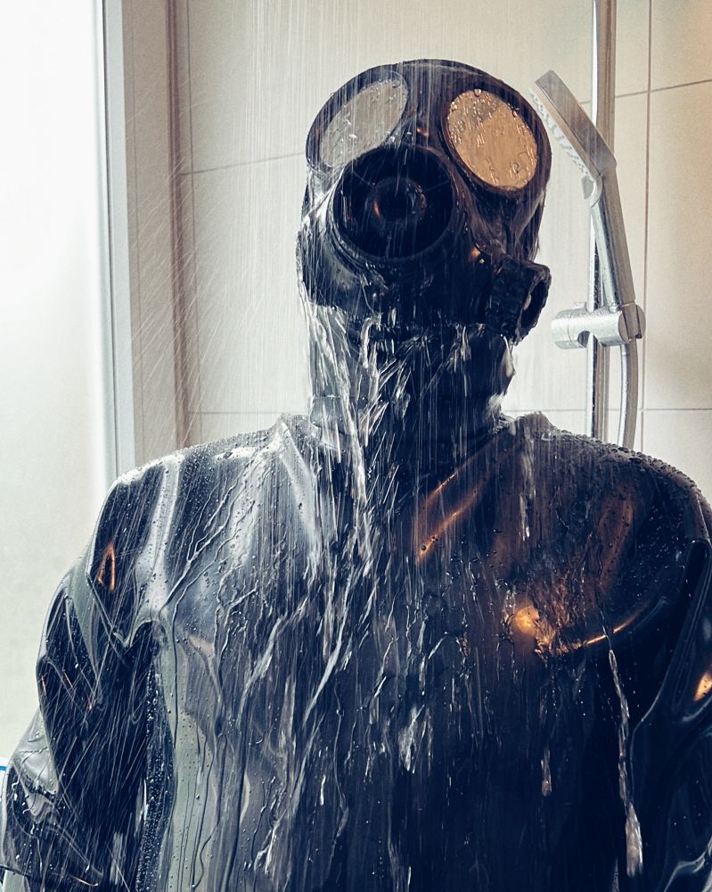 A person in a black heavy rubber suit and a gasmask is taking a shower.
