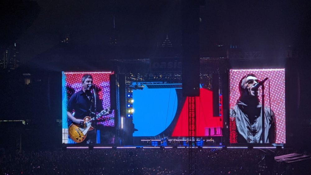 Imagen de un escenario de un concierto con dos pantallas a sus extremos. En las pantallas se proyectan imágenes de Noel y Liam Gallagher del grupo Oasis. 