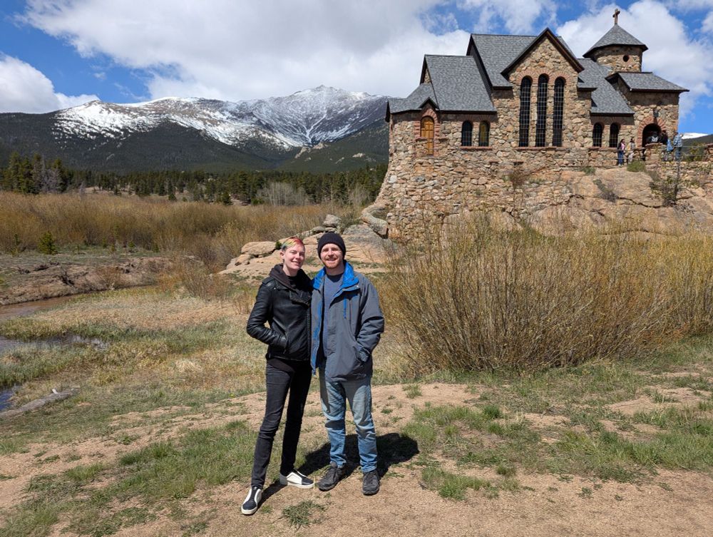 Apex stands smiling with their partner in a black leather jacket with their hands in their pockets, dark gray jeans, and black and white leather sneakers. Their partner is a white masculine individual wearing a black beanie, blue and gray coat, blue jeans, and dark gray hiking shoes. In the background there is a stone chapel and the Rocky Mountains