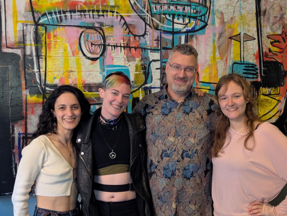 A photo of Apex, two of their femme metamours, and our masculine mutual partner Jeff dressed casually for his birthday outing. There is a busy, contemporary art piece in the background