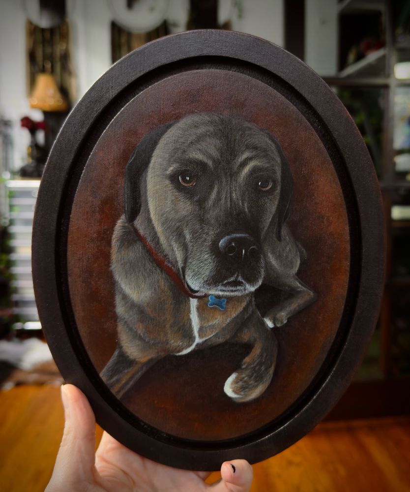 Hand holding a wood plaque painted with acrylics of a dog portrait. The dog is a Boxer Rottweiler mix with the brown and black brindle pattern. She is in the lying position with one paw curled under and her head up and looking forward. 