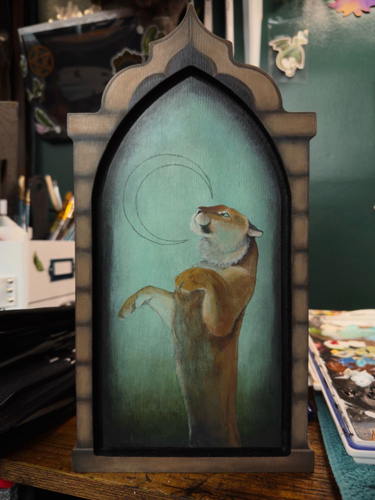 Sitting on a desk is a window shaped wood plaque with a painting started. The painting depicts a beige stoned window around the edge of the plaque. On the center of the plaque is a tiger (without stripes) standing up reaching its front legs up towards a pencil outline of a crescent moon. The background transitions from green at the bottom to a teal and blue at the top. This piece is still a work in progress so colors and blending of the tiger is not smooth and uniform yet. 