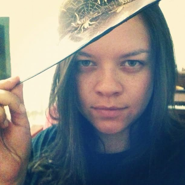 selfie própria - mulher branca com a expressão facial padrão de toda feia, cabelo escuro liso caindo sobre o ombro e um chapéu de bruxa de tule rosa com teias de aranha douradas 