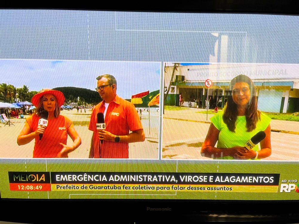 Foto da tv em grande do jornal local da hora do almoço. A tela dividida em duas partes. Do lado esquerdo, dois apresentadores - uma mulher e um homem- vestidos em roupas num tom reluzente de laranja, mal cortadas, com um chapéu de cata ovo ridículo na mesma cor. Do lado direito, uma coitada usando o mesmo uniforme em verde limão, com um caimento péssimo no tronco, braços e pescoço. A saturação deixa o vídeo horrível, as cores no entorno apagadas e a pele das pessoas parecendo pós uso de Dove barro