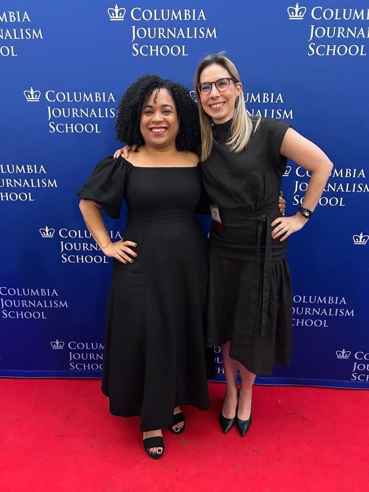 Two female journalists wearing black, long dresses, standing in front of a Columbia Journalism School backdrop. 