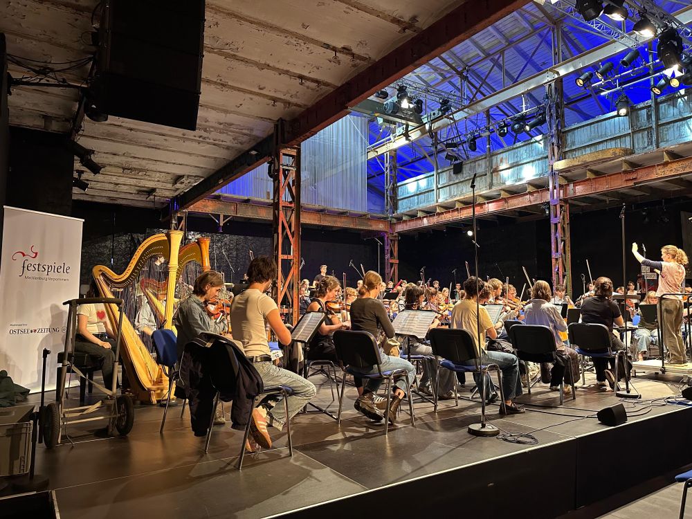 Side view of orchestra rehearsal from back of the 1st violin section, you can see two harpists on the left of the photo and the conductor with an arm raised in the right hand side.
