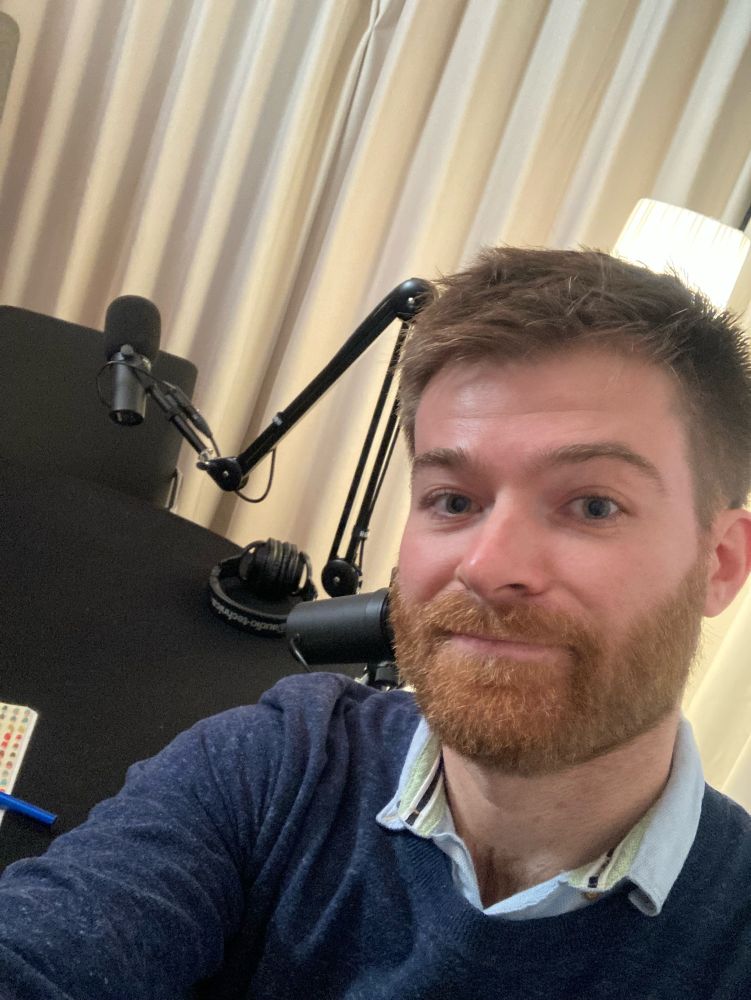 A picture of a man - me - sitting with two studio microphones and headphones behind him on a table.