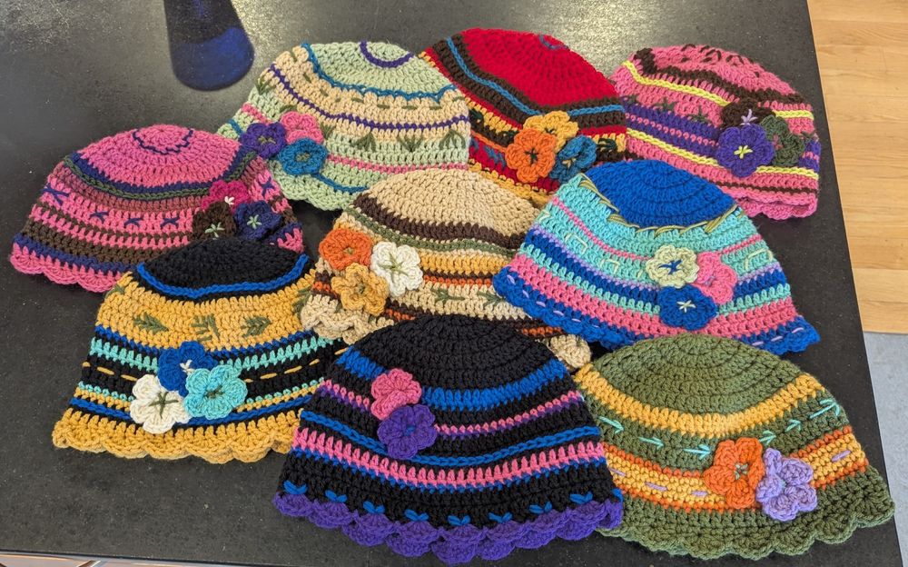 Nine colorful crocheted hats laid out on a black countertop. Each has stripes, crocheted flowers, various embroidery, and scalloped bottom edge. 