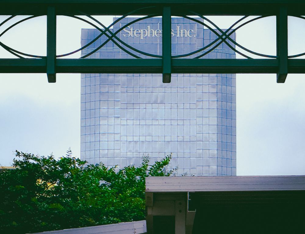 A skyscraper in Little Rock, Arkansas, that has a gray blue reflective glass and his framed thigh, green trees, and the fancy green railing of a parking deck.