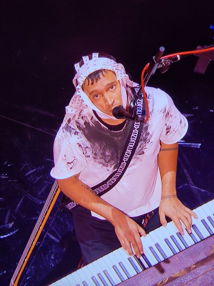 Photo from above of a man (Tyler Joseph) at the keys of a piano while singing into a microphone. He is wearing a white head piece that wraps around his throat and sides of his face with fastener stripes falling back and framing.