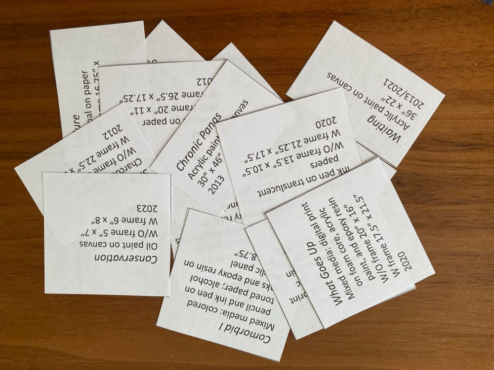 A bunch of little paper slips with info about the artworks for the upcoming show.  The text includes the title, media, size, and date completed.  The paper slips are piled up and disorganized. 