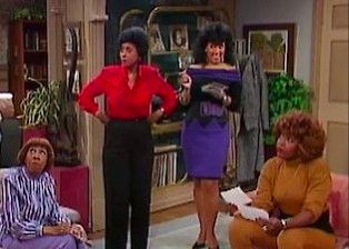 Four women are gathered in a cozy living room. One woman in a red blouse and black pants stands with her hands on her hips. Next to her, another woman in a purple off-the-shoulder dress and black belt holds papers and smiles. A woman in a mustard-yellow outfit sits on a chair holding sheets of paper, looking toward the others. Another woman, dressed in a striped blouse and purple pants, sits on the couch glancing upward. Bookshelves, plants, and framed artwork decorate the room.
