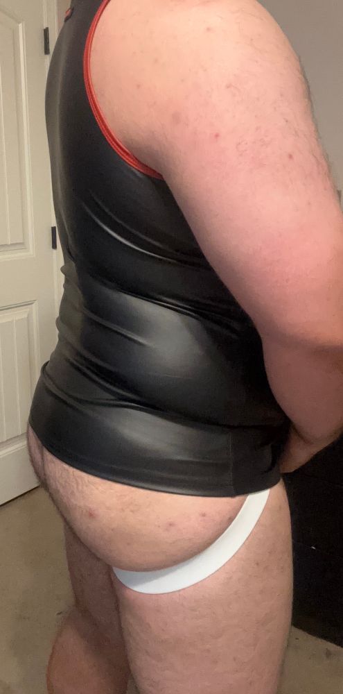 Me, wearing a black tank top with red piping and a white jockstrap. The tank top is hiding the top part of my jock. I’m turned 3/4 away from the camera, showing off part of my side and ass