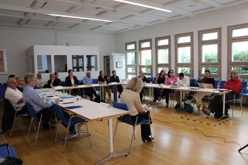 Menschen sitzen an u-förmig aufgestellten Tischen und hören einem Vortrag zu.