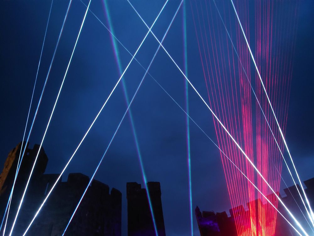 Lasers criss-crossing a dark blue evening sky with castle towers in silhouette.