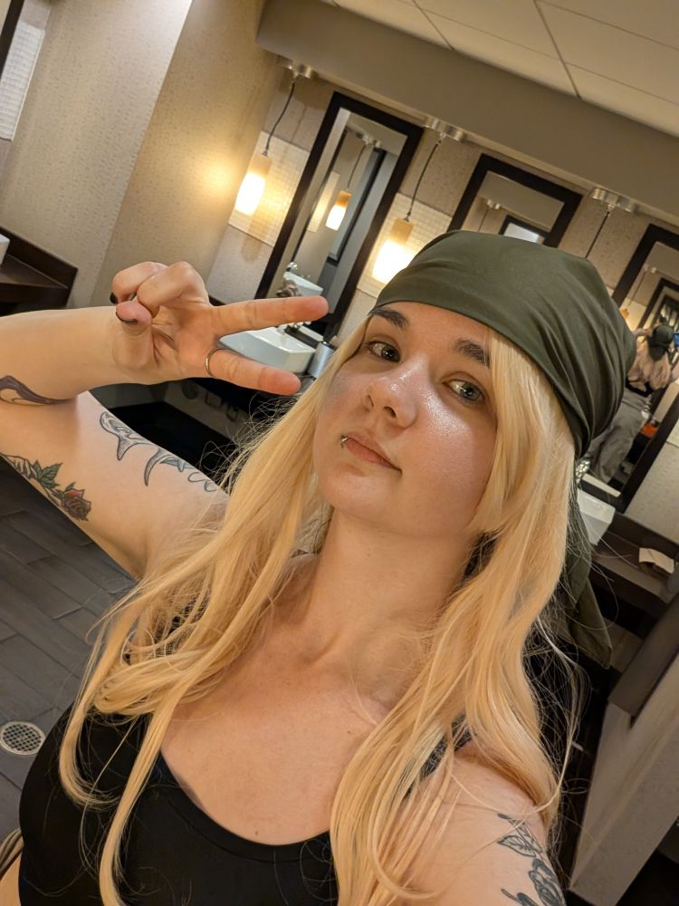 A selfie of my in the bathroom. I have on a blonde wig and green bandana and a black sports bra. I'm holding up a peace sign by my face.