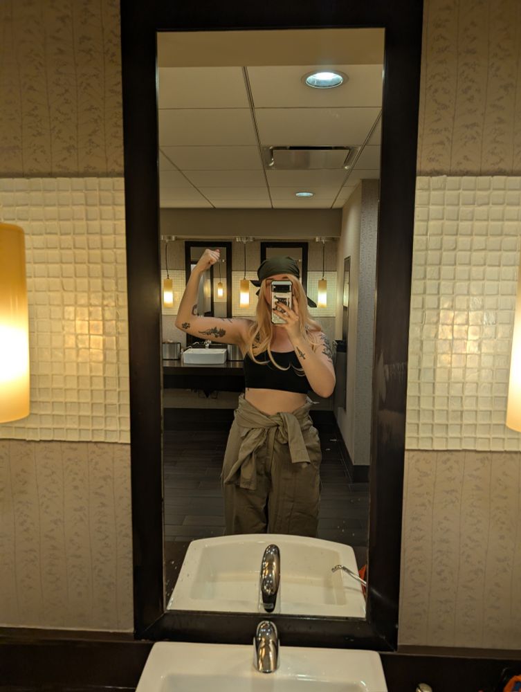 A photo of me in an empty bathroom posing in front of a mirror. My phone is blocking my face, but you can see the long blonde hair and green bandana on my head still. I have my left arm raised up as if I'm flexing it. I have on a black sports bra and a tan pair of coveralls with the sleeves tied around my waist.