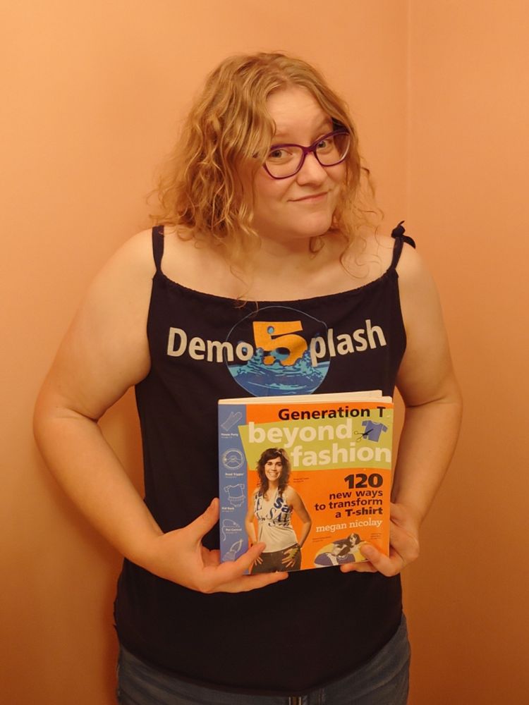 A woman wearing a tank top made from a Demosplash 2015 shirt, with the front design facing the camera, and holding a copy of Megan Nicolay 's Generation T: Beyond Fashion.