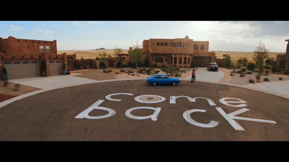 Screenshot from the Apple TV show, Pluribus. An drone shot of a cul-de-sac with the words "come back" written on the ground, with two lesbians in the background hugging. This image represents my appeal to people on Bluesky to come back to Twitter because I miss them and I cannot get used to this website.