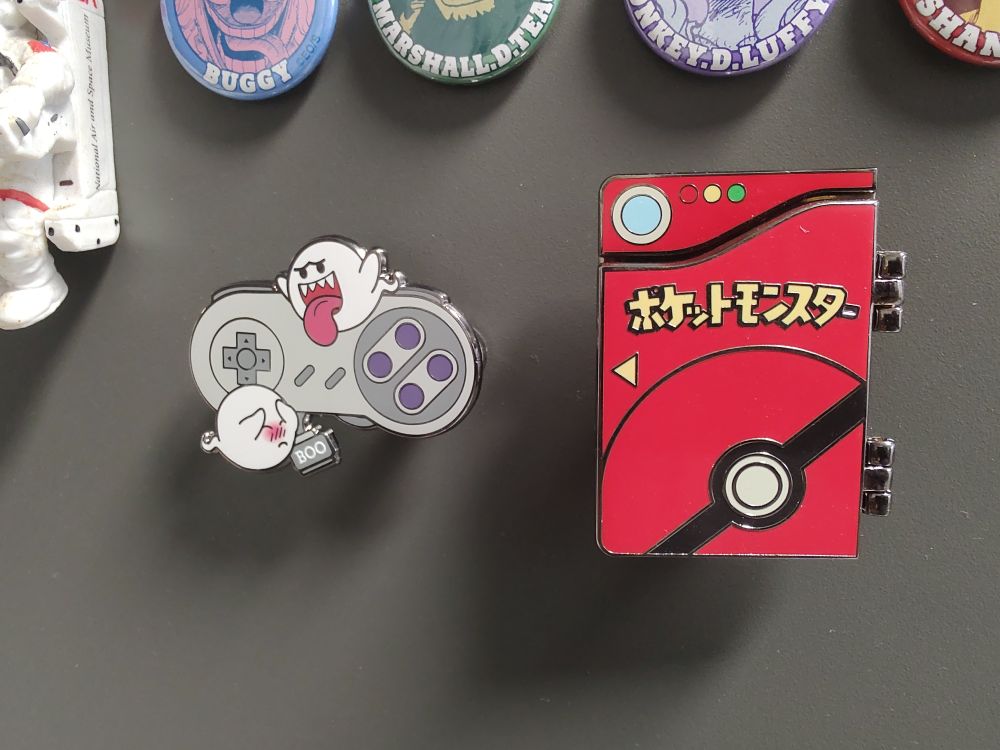 Foto de dois pins. Um deles representa um controle de super Nintendo com dois Boos voando ao redor dele e o outro é uma Pokedex da primeira geração de Pokémon 