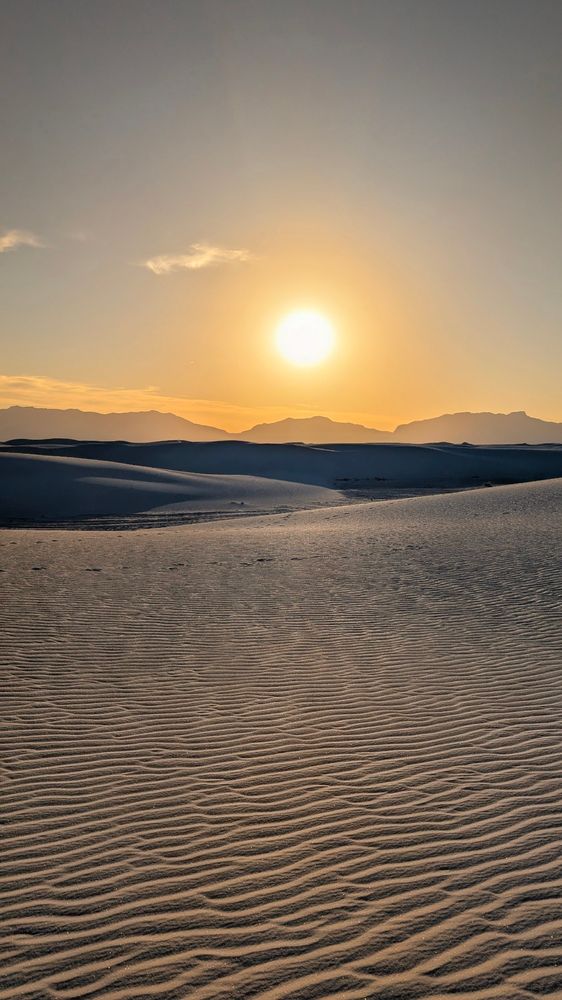 The sun sets about mountains on the horizon beyond rippled sand. 