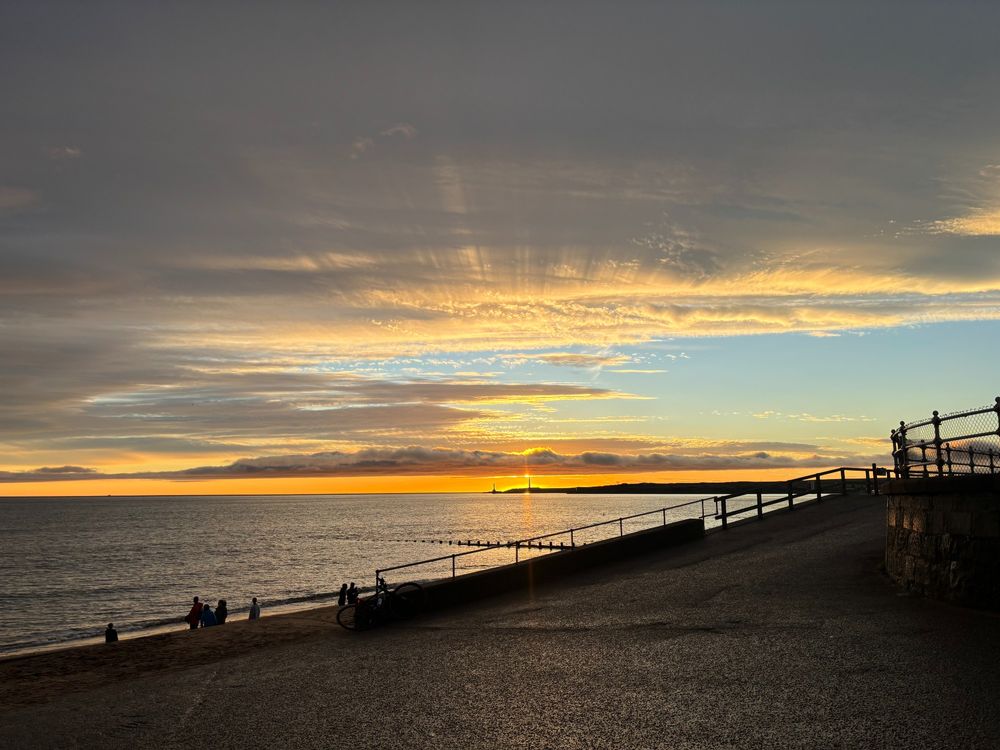 Mid-winter sunrise over the north sea. A lighthouse is silhouetted by the sun. Rays of light illuminate low cloud. A patch of blue sky is visible in the middle right of the image.
