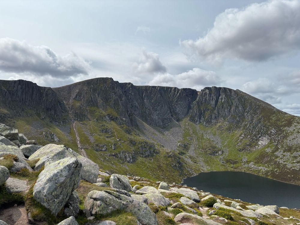 The coire of Lochnagar. The loch is in the bottom right corner of the image. A Wide cliff filled with gullies and buttresses covers the middle of the image. White cumulus nimbus clouds cover the top third.