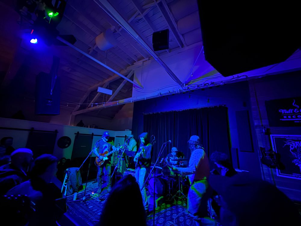 Rock band performing on stage underneath blue-ish lighting, in a warehouse setting