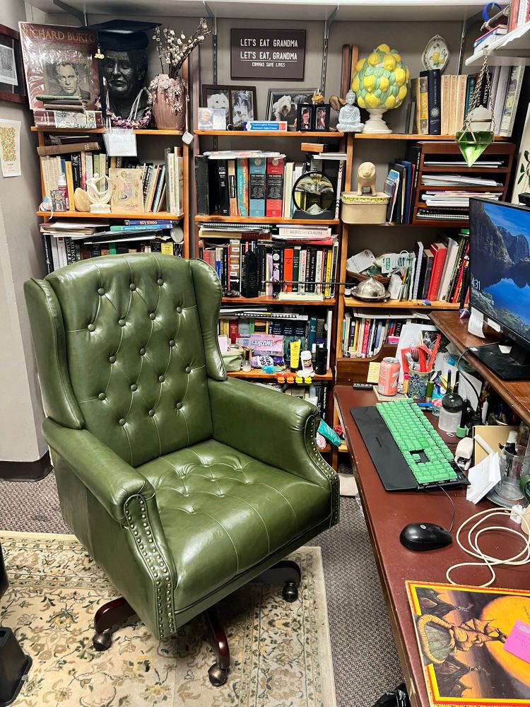 Olive green wingback office chair in front of busy bookcases and a desk made out of an old upright piano. 