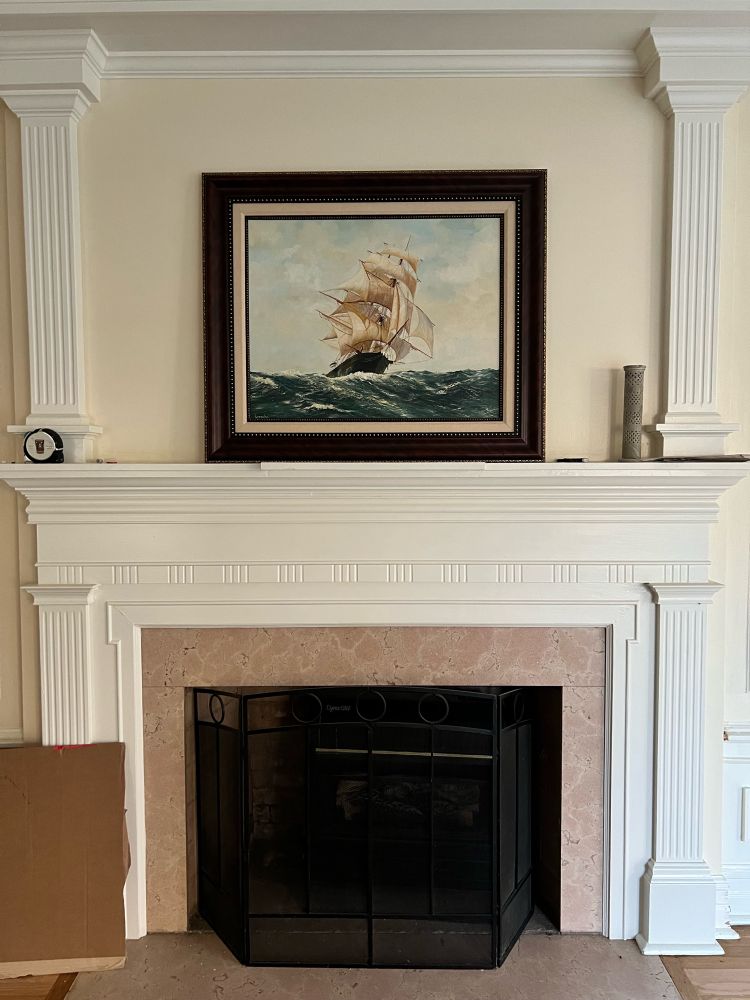 Framed oil painting of a sailing ship under full sail sitting on a mantle over a pink marble fireplace. 